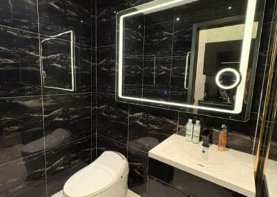Modern black-tiled bathroom with illuminated mirror and white toilet.